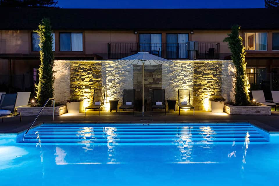 Illuminated resort pool at night with stone features, lounge chairs, and building backdrop