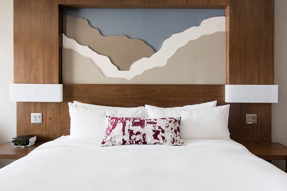 Modern hotel bedroom with wooden headboard, white bedding, burgundy accent pillow, and minimalist artwork