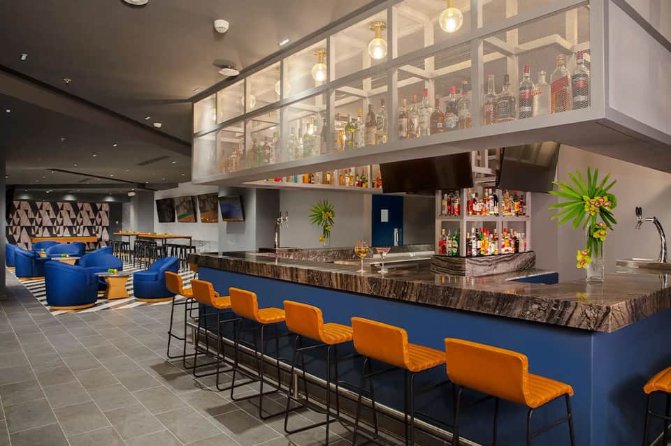 Modern hotel bar with blue counter, orange seating, and illuminated bottle display
