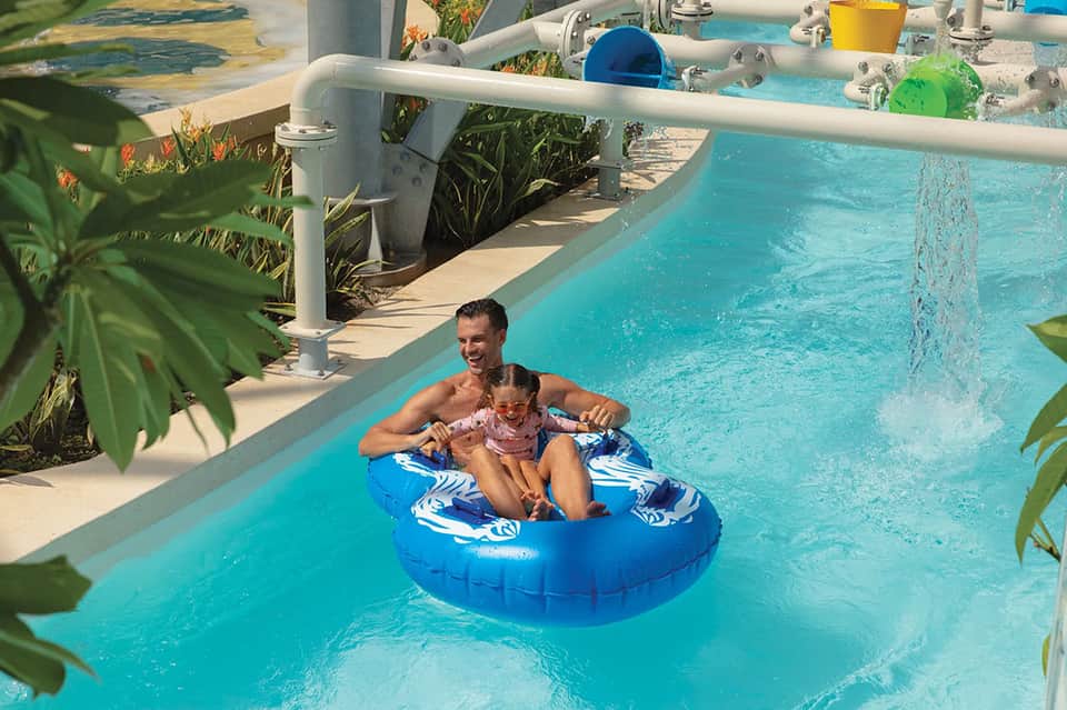 Family enjoying lazy river pool float at tropical resort with turquoise water and lush vegetation