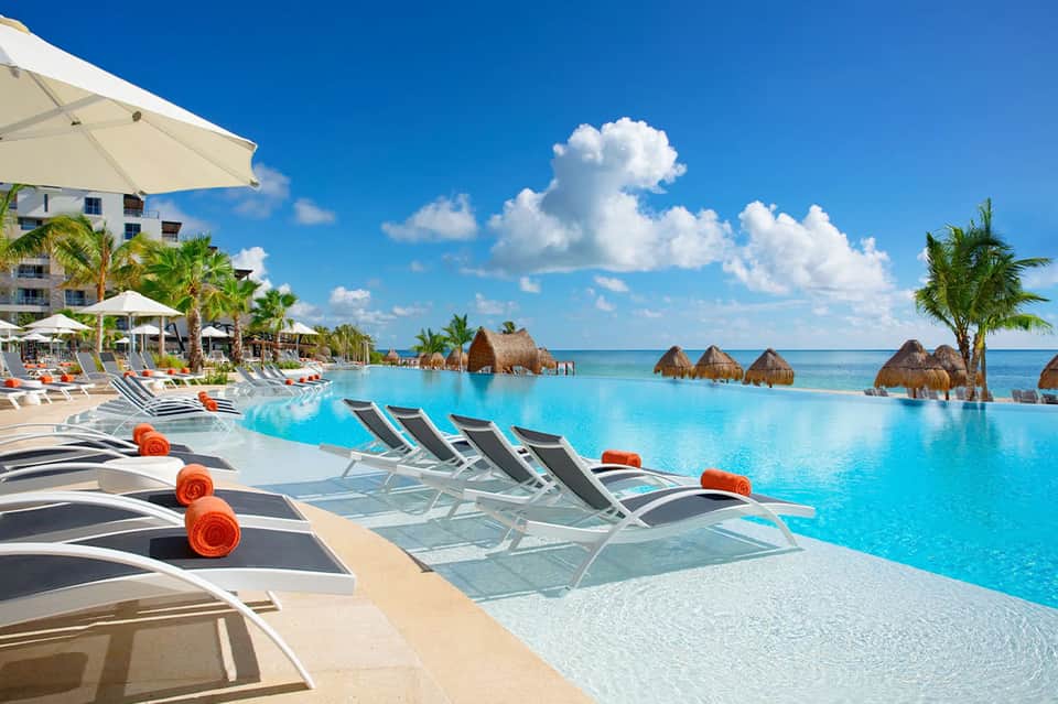 Large resort pool with turquoise water, tiki huts, palm trees, ocean view, lounge chairs with towels