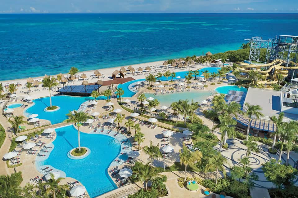 Aerial resort view with blue pools, beachfront, palm trees, and turquoise ocean waters