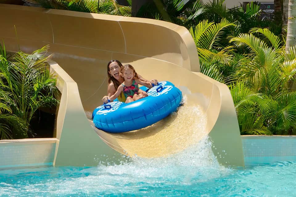 Woman and child sliding down water slide into turquoise pool with tropical vegetation surrounding