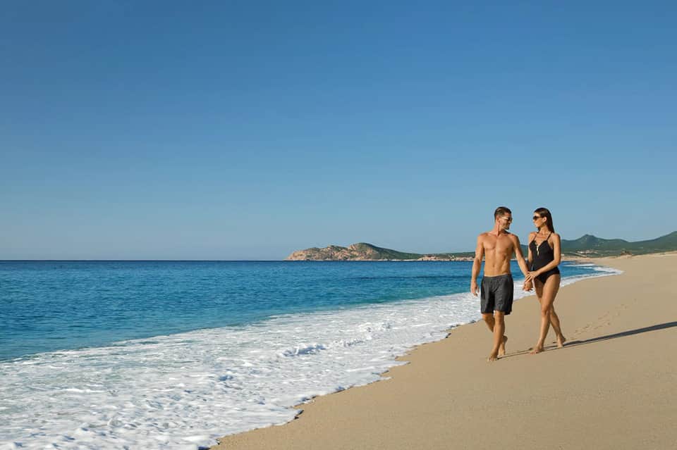 Couple walking along sandy beach with turquoise ocean waves and coastal mountains