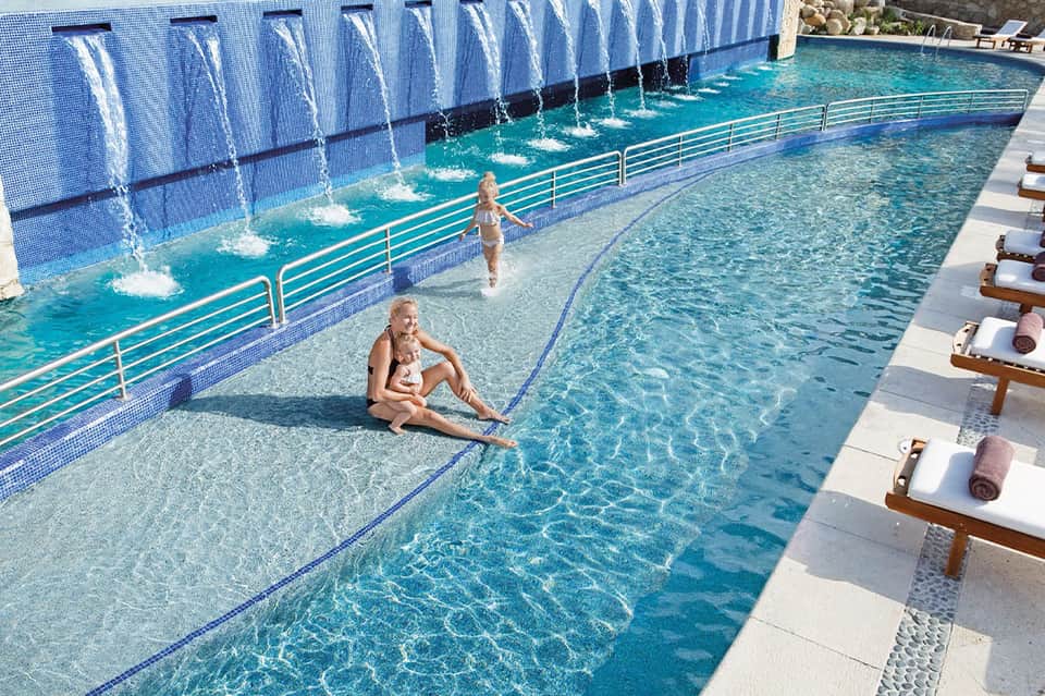 Resort swimming pool with waterfall feature wall, guests in shallow water, and lounge chairs with towels lining deck