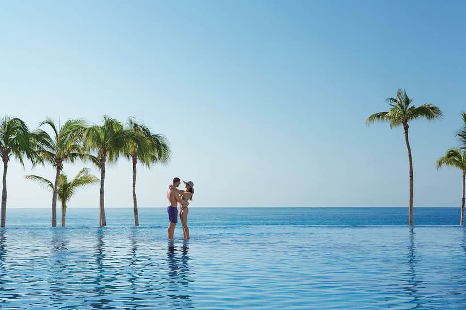 Infinity pool overlooking ocean with couple in water, palm trees, and clear blue horizon