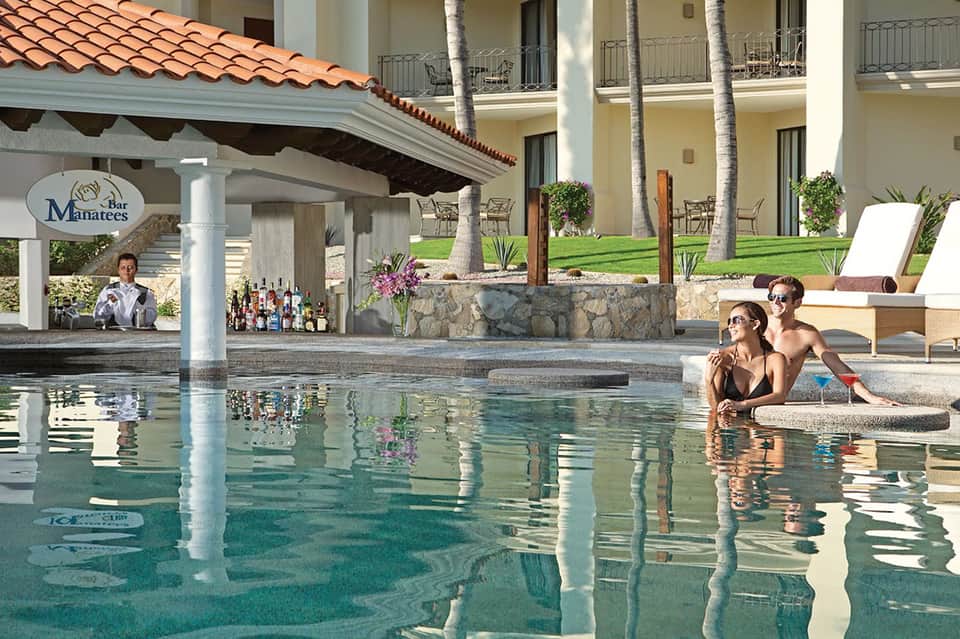 Resort pool bar with bartender, guests swimming, and Spanish-style architecture overhead