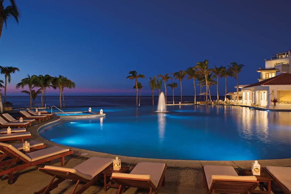 Luxury infinity pool at dusk with illuminated water, loungers, palms, and ocean view