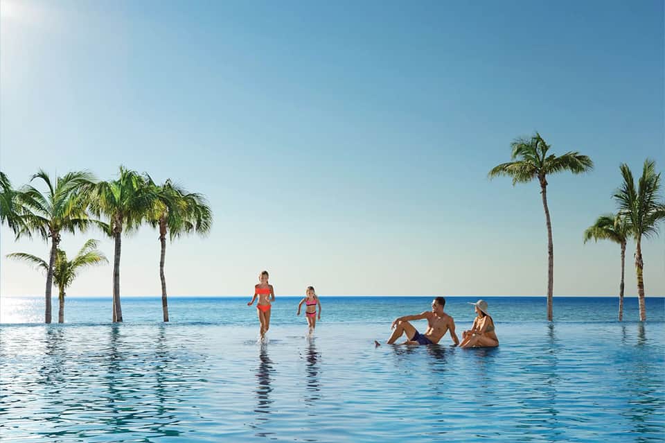 Infinity pool with guests swimming and palm trees overlooking a blue ocean horizon