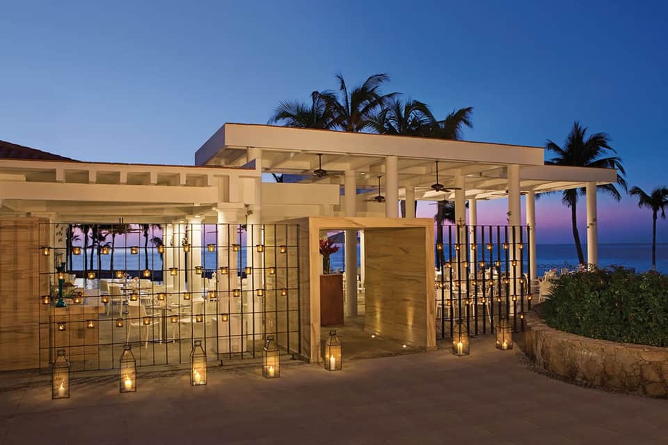 Modern beachfront structure with glass walls, white columns, and palm trees at dusk