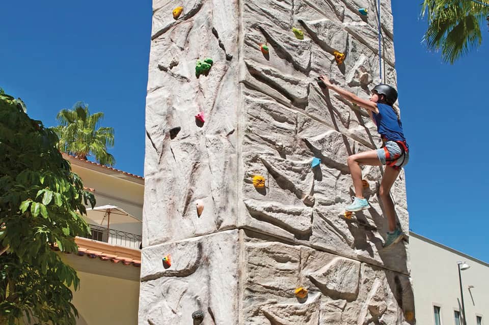 Child rock climbing an indoor climbing wall with colorful handholds, harness, and safety equipment in resort