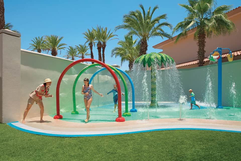 Family enjoying colorful water play arches in shallow splash pad surrounded by palm trees and resort grounds