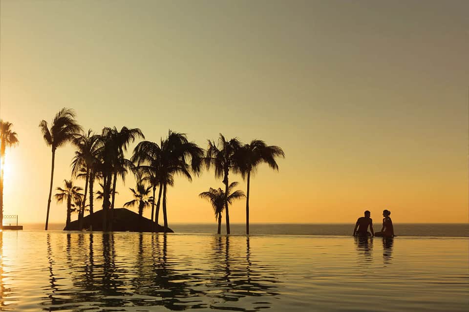 Sunset reflection in infinity pool with silhouetted palm trees and two people wading