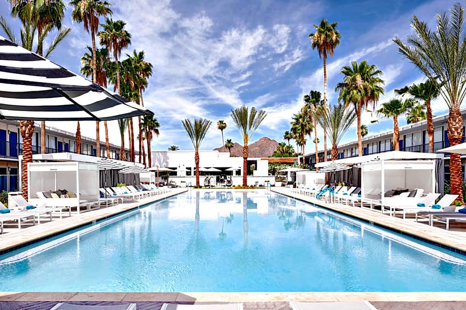 Resort pool surrounded by white lounge chairs, palm trees, and cabanas with striped umbrellas under blue sky