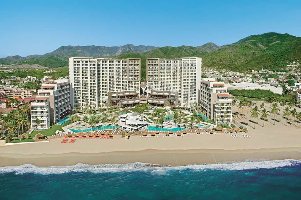 Beachfront resort aerial view with multiple pools, palm trees, sandy beach, and green mountains