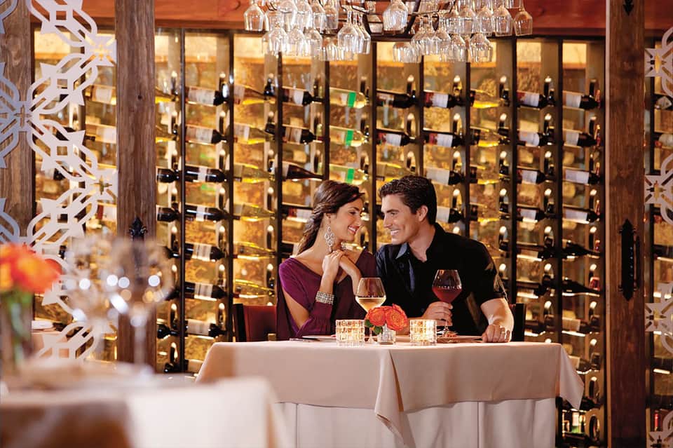 Couple dining at an elegant restaurant table with illuminated wine cellar wall in background