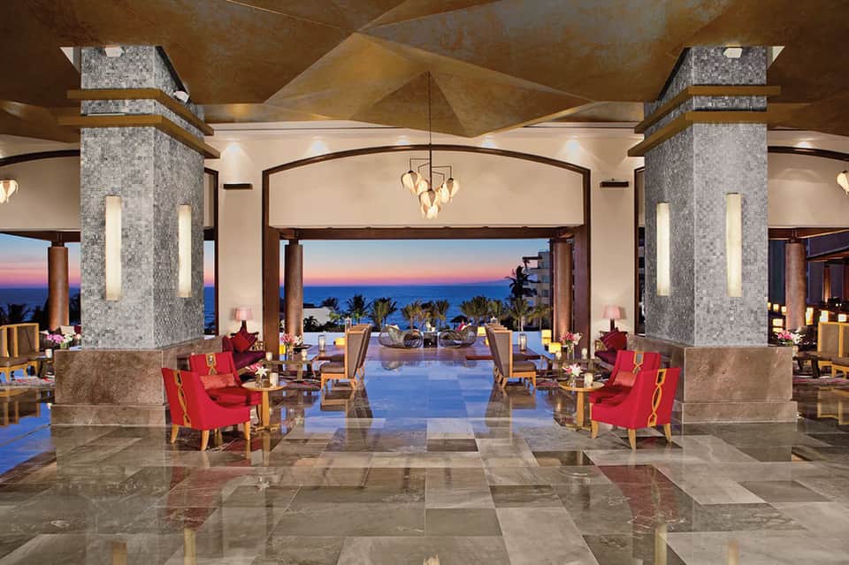 Luxury resort lobby at dusk with marble floors, red accent chairs, soaring ceiling, and ocean view