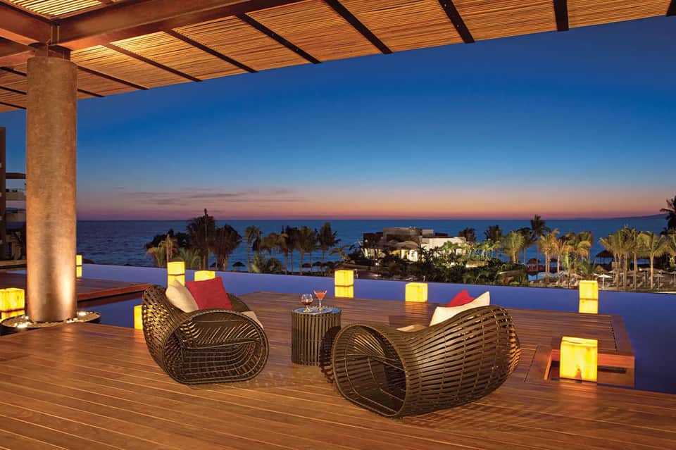 Waterfront deck at dusk with woven seating, illuminated cubes, and ocean sunset view