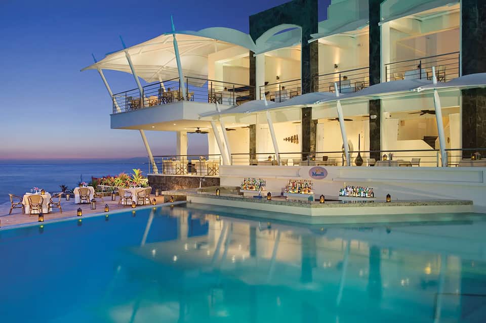 Luxury resort pool at dusk with modern architecture, illuminated bar, and ocean views