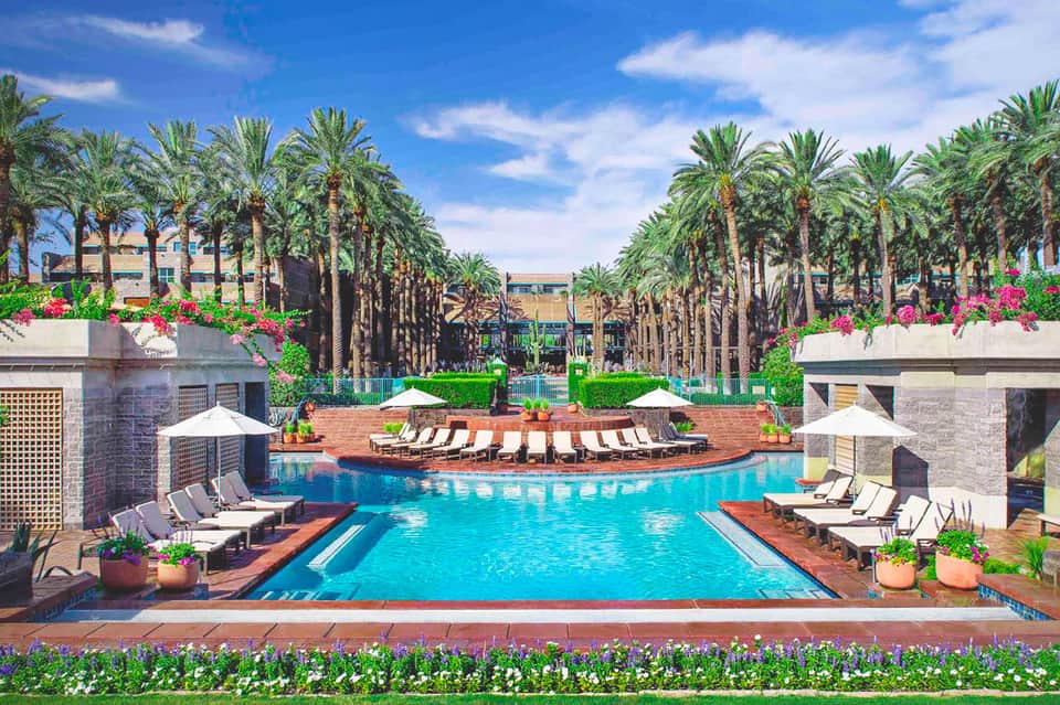 Luxurious resort pool with crystal blue water, palm tree lined courtyard, brick deck, lounge chairs, and vibrant pink flowers