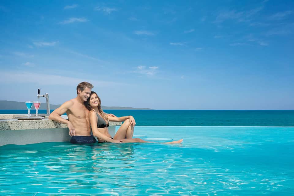 Couple in infinity pool overlooking calm turquoise ocean horizon with colorful cocktails nearby