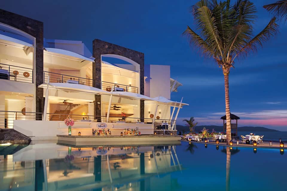 Luxury modern villa lit at twilight with infinity pool, illuminated interior, and palm tree silhouette