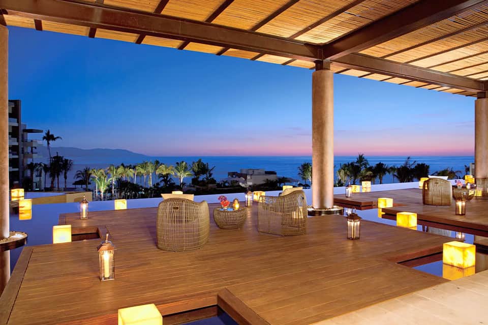 Twilight ocean view lounge with woven seating, palm trees, and candlelit ambiance