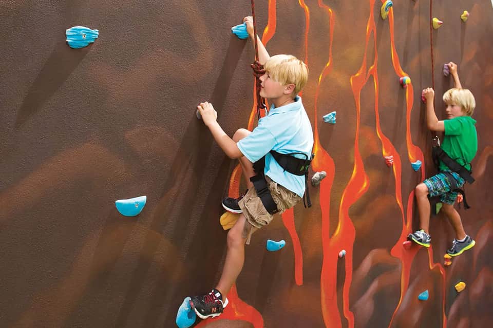 Children climbing indoor rock wall with orange and yellow holds and safety equipment