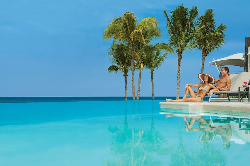 Couple relaxing by infinity pool overlooking ocean with palm trees and clear blue sky