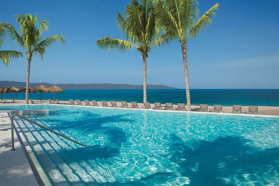 Resort pool with turquoise water, palm trees, tiki umbrellas, lounge chairs, and ocean coastline backdrop