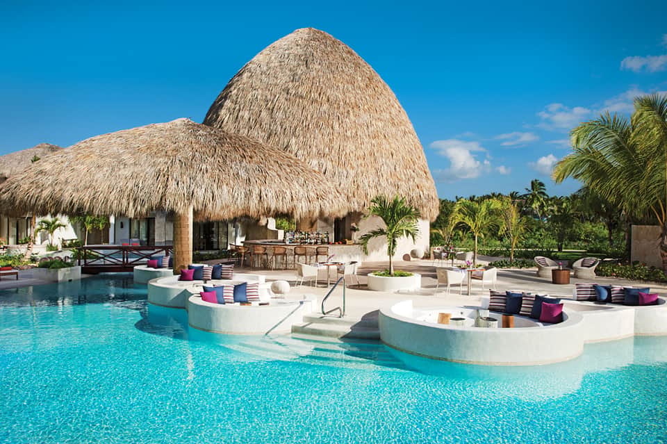 Luxury resort pool with thatched bungalows, in-pool lounging platforms, and palm trees