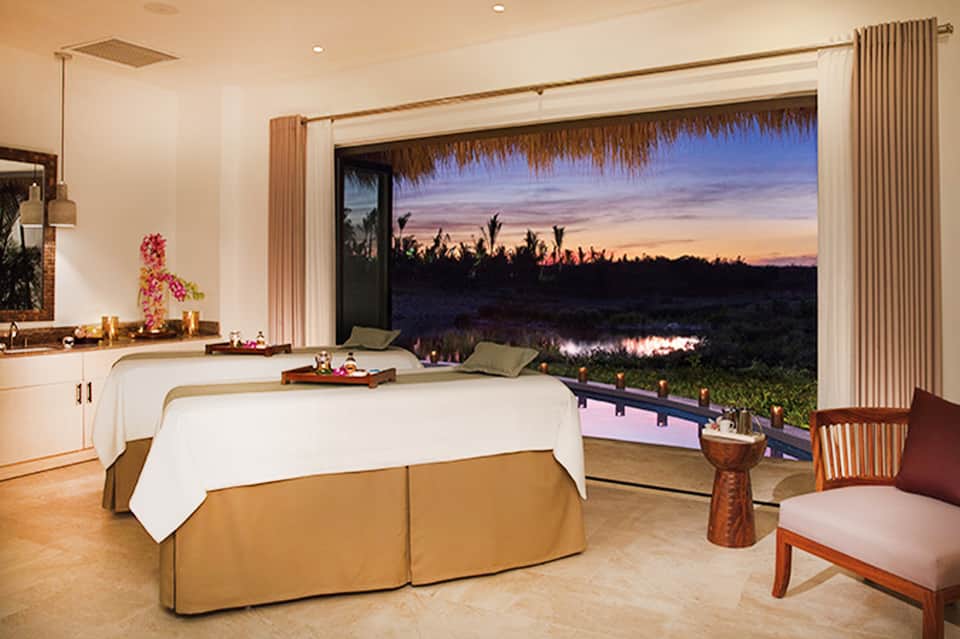 Spa treatment room with two massage beds overlooking sunset wetlands and palm trees