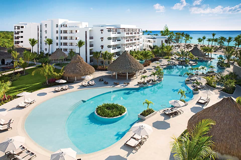 Aerial view of beachfront resort with curved pool, palm trees, thatched structures, and ocean