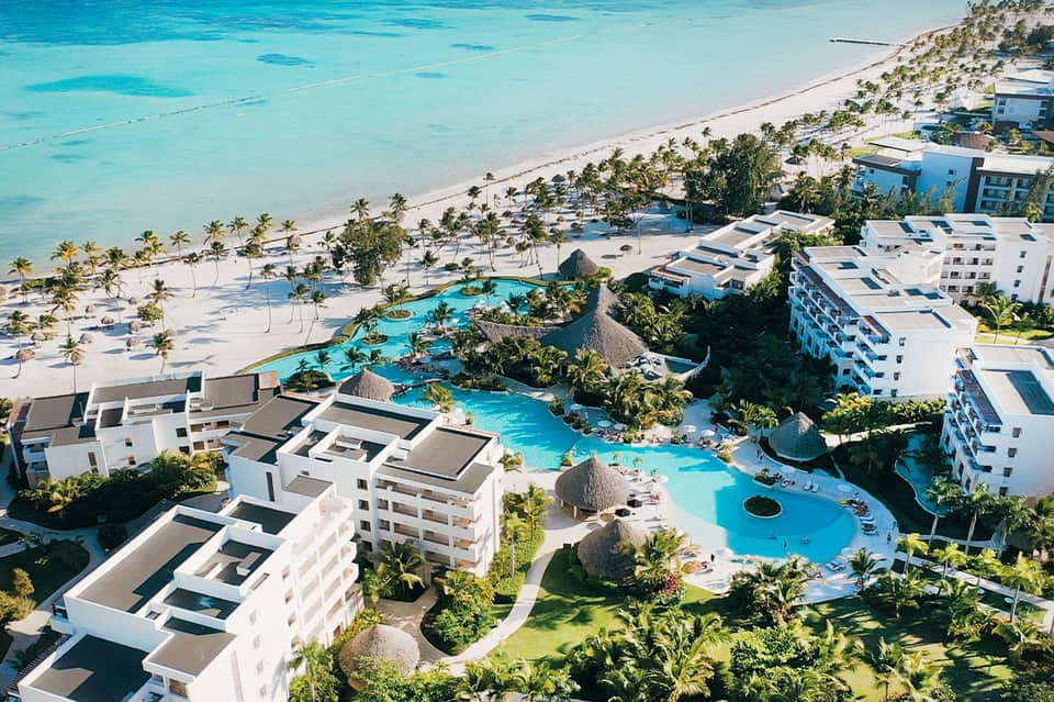 Aerial view of beachfront resort with turquoise pools, white buildings, palm trees, and pristine beach