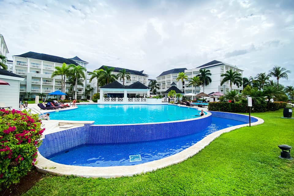 Resort swimming pool with blue water, white buildings, palm trees, and manicured grounds