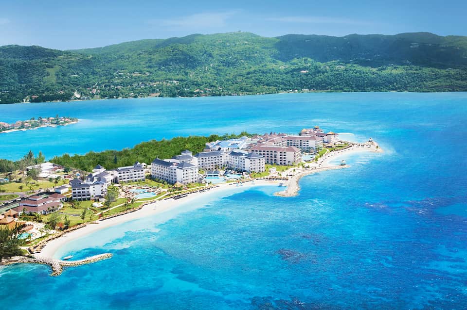 Aerial view of beachfront resort with white buildings, sandy beach, turquoise lagoon, and lush green mountains