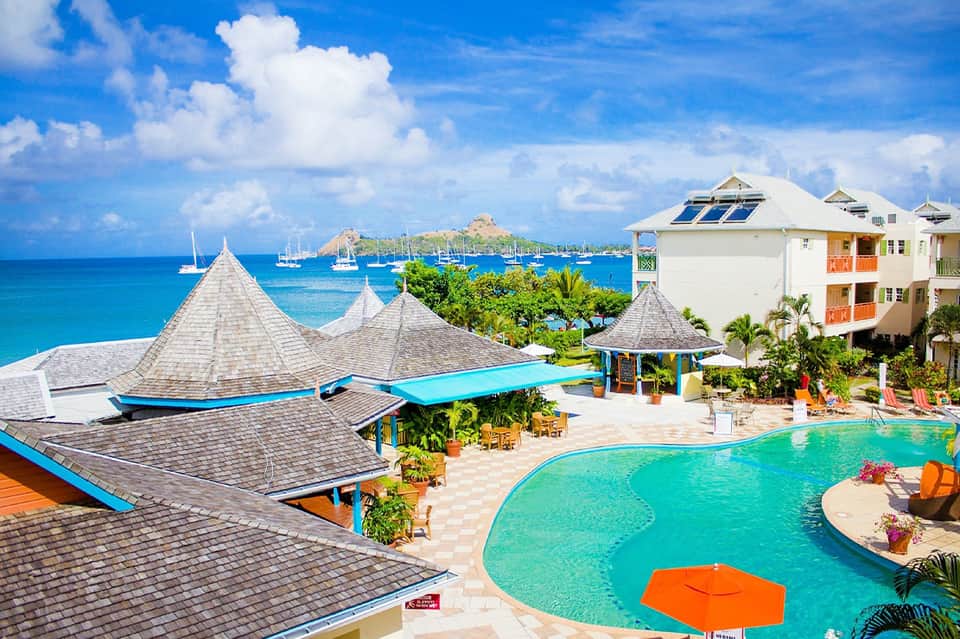 Tropical resort pool with teal water, thatched-roof pavilions, and colorful lounge umbrellas overlooking sailboats