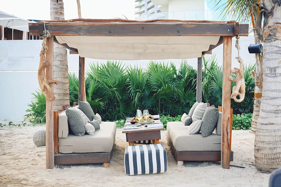 Wooden beach cabana with cream cushioned seating, striped ottoman, and ocean view