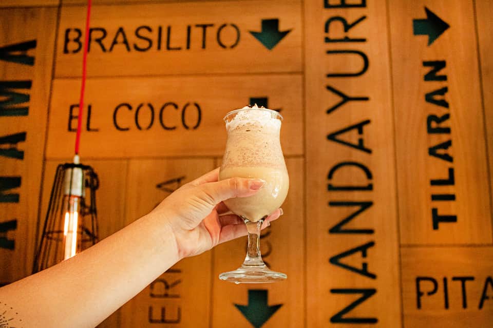 Hand holding creamy tropical cocktail against wooden menu board with drink names