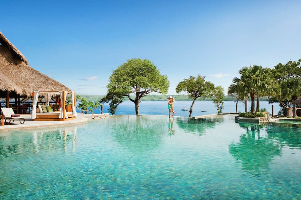 Infinity pool overlooking tropical lake with thatched cabanas, palm trees, and mountain views