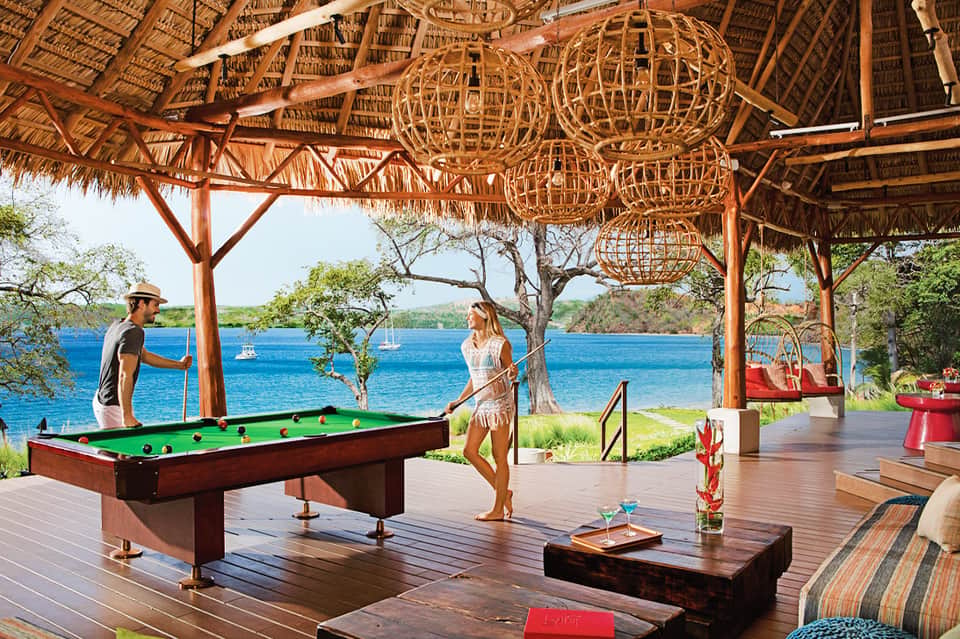 Couple playing billiards on covered wooden deck overlooking blue water and boats with tiki decorations