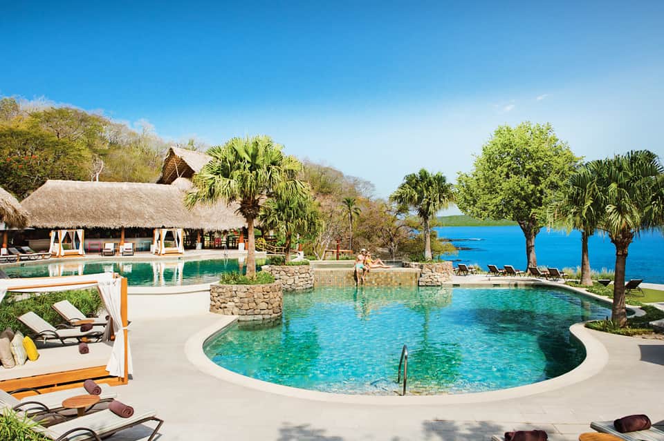 Luxury resort pool with thatched-roof buildings, palm trees, and turquoise lagoon view