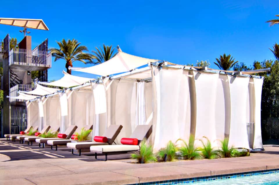Luxury poolside cabanas with white curtains, loungers with red cushions, and palm trees under clear blue sky