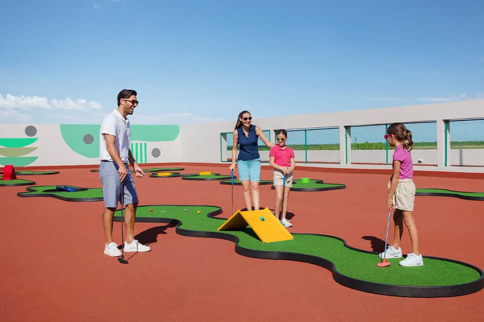 Family playing miniature golf on colorful course with red surface and green obstacles