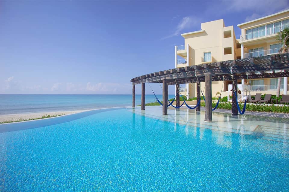 Infinity pool overlooking pristine beach and ocean with wooden pergola and resort buildings