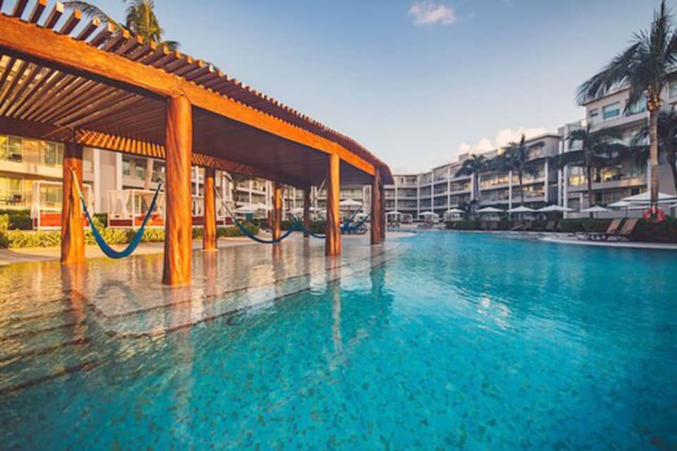 Resort pool with wooden pergola and hammocks, flanked by palm trees and multi-story buildings