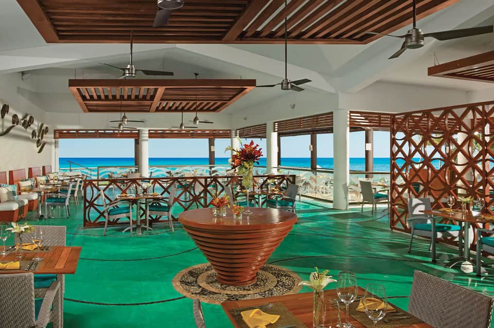 Beachfront restaurant with green floor, wooden ceiling, turquoise ocean views, and elegant table settings.