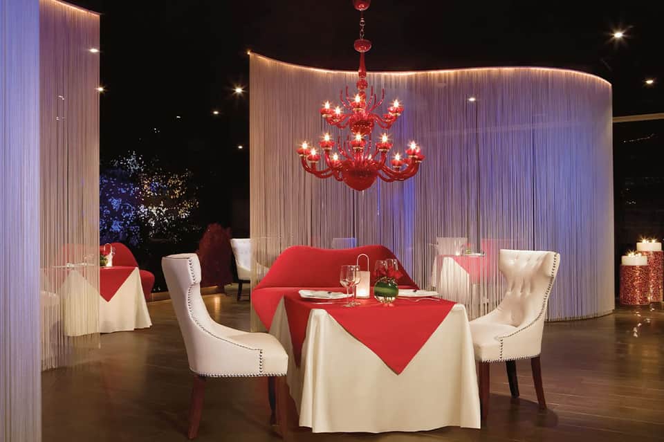 Elegant fine dining table setting with red tablecloth, white chairs, red chandelier, and ambient purple lighting.