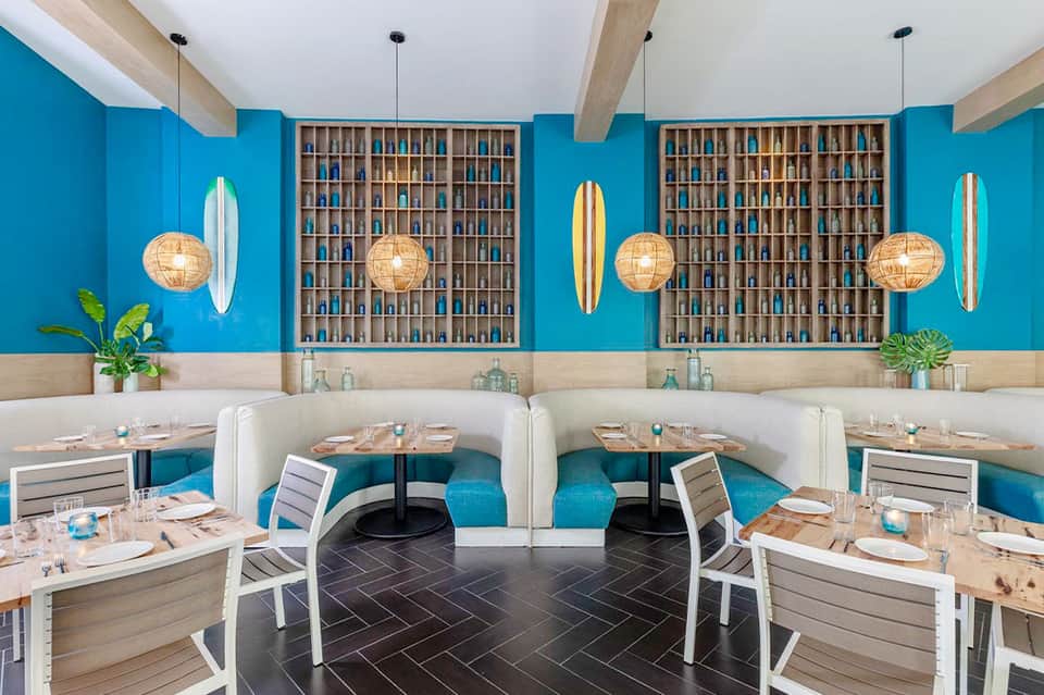 Contemporary restaurant interior with bright blue walls, wooden shelving, rattan pendant lights, and turquoise booth seating