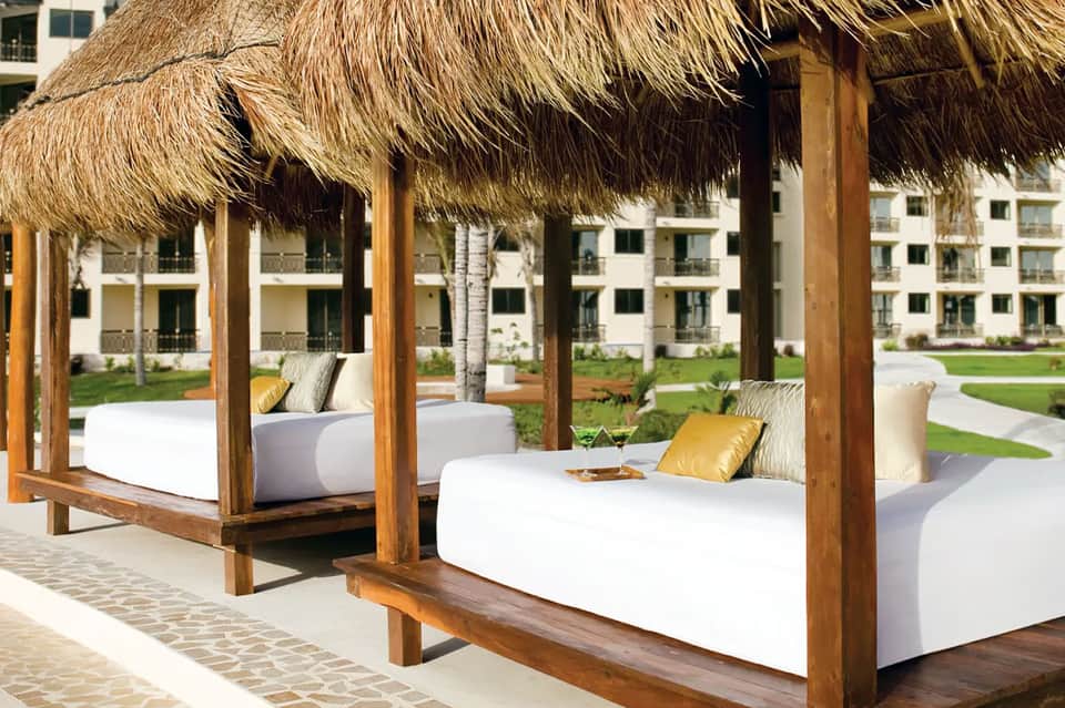 Thatched-roof lounge cabanas with white cushioned daybeds overlooking tropical resort grounds